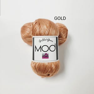 May include: A skein of metallic gold yarn with a white paper label featuring the text "Crochet Yarns MOD" and a small logo. The yarn has a shiny, smooth texture and is wound into a rounded shape, displaying a consistent warm, reflective color.
