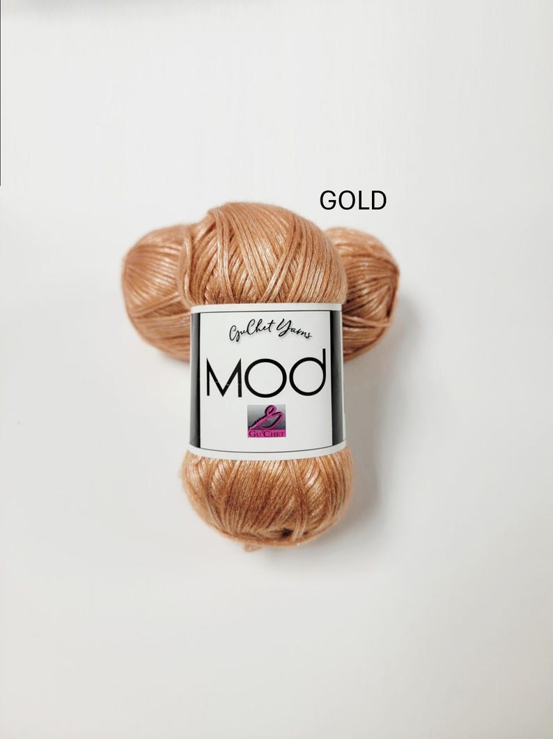 May include: A skein of metallic gold yarn with a white paper label featuring the text "Crochet Yarns MOD" and a small logo. The yarn has a shiny, smooth texture and is wound into a rounded shape, displaying a consistent warm, reflective color.