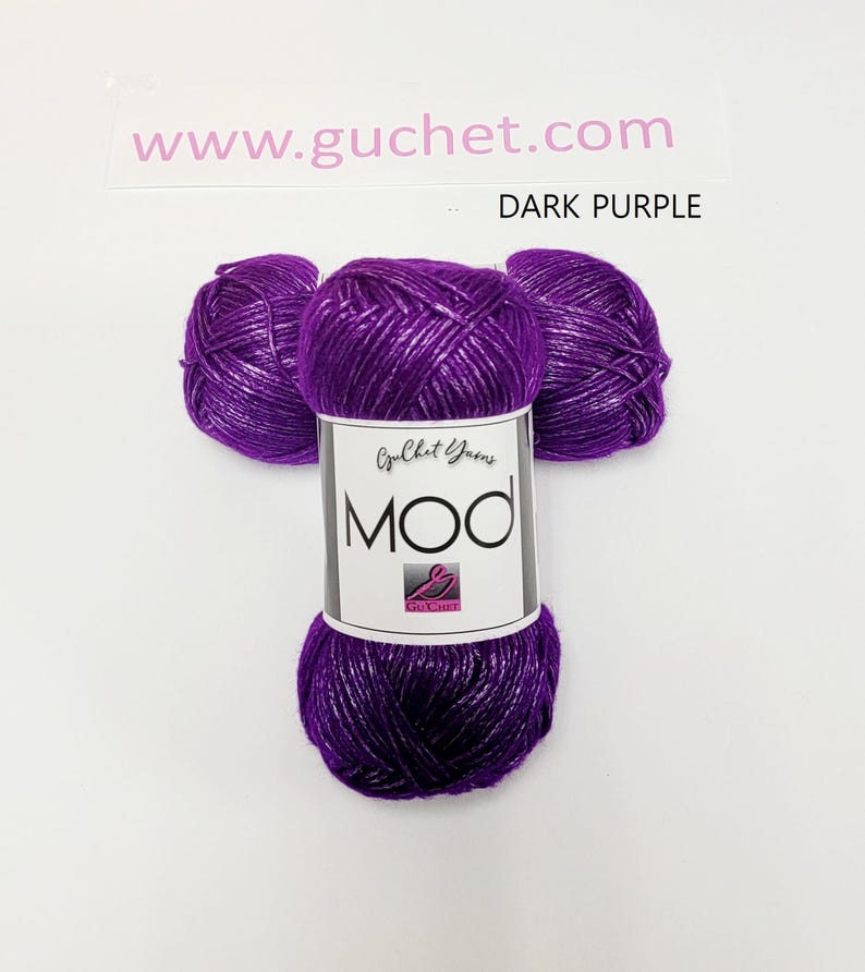 May include: Three skeins of dark purple yarn with a metallic sheen, arranged in a cluster. A white paper label wraps around the center, featuring the text Guchet Yarns, MOD, and the website www.guchet.com with the color label DARK PURPLE.