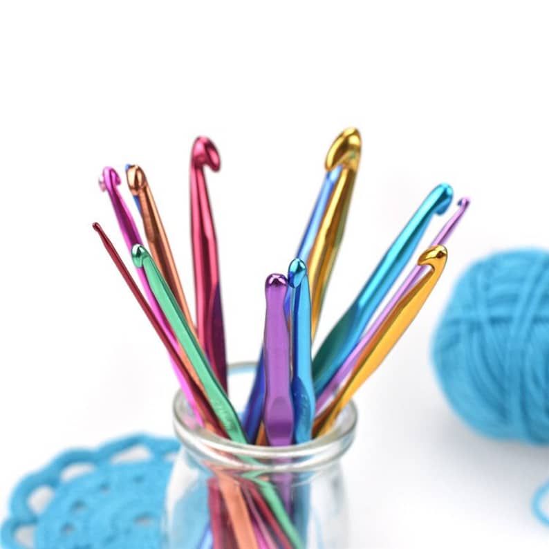 Op de afbeelding: Een verzameling kleurrijke haaknaalden in een heldere glazen pot. De haken hebben verschillende metallic kleuren, waaronder roze, groen, blauw en goud. Een bolletje blauw garen en een blauw gehaakt kleedje zijn op de achtergrond.