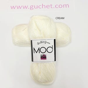 May include: A skein of cream-colored yarn with a white label featuring the text Guchet Yarns MOD and a small pink logo. The yarn has a soft, slightly shiny texture. The text www.guchet.com is visible above the yarn, and the word CREAM is to the right.