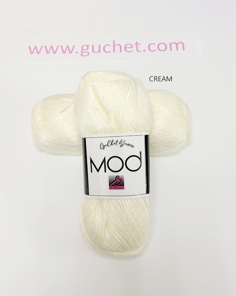 May include: A skein of cream-colored yarn with a white label featuring the text Guchet Yarns MOD and a small pink logo. The yarn has a soft, slightly shiny texture. The text www.guchet.com is visible above the yarn, and the word CREAM is to the right.