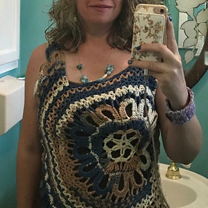 May include: A woman wearing a crocheted top with a blue, brown, and white color scheme. The top has a circular design with a floral pattern in the center. The woman is wearing white shorts and has a beaded necklace.