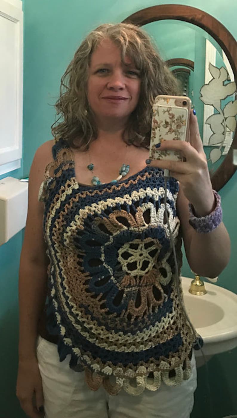 May include: A woman wearing a crocheted top with a blue, brown, and white color scheme. The top has a circular design with a floral pattern in the center. The woman is wearing white shorts and has a beaded necklace.