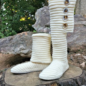 May include: A pair of white knitted slipper boots with button accents. The boots are sitting on a tree stump in a natural setting.