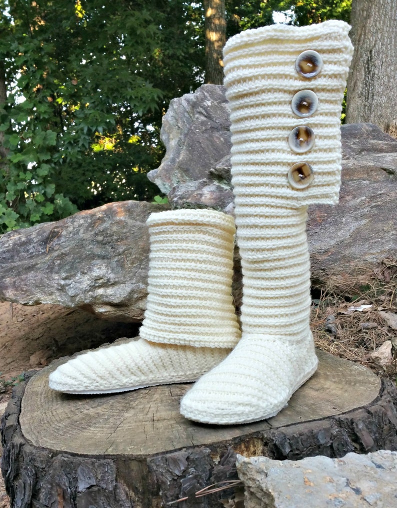 May include: A pair of white knitted slipper boots with button accents. The boots are sitting on a tree stump in a natural setting.