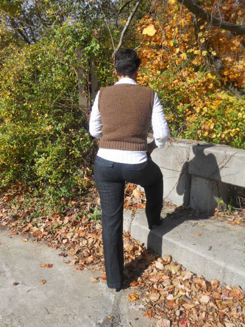 May include: A person wearing a brown knitted vest with a white shirt and black pants. The person is standing on a concrete surface with fallen leaves.