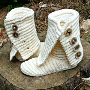 May include: A pair of white crocheted slipper boots with brown button accents. The boots have a fold-over cuff and are made with a soft, textured yarn.