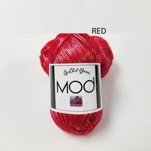 May include: A ball of bright red yarn with a metallic sheen. The yarn is wrapped in a white paper label featuring the text "Guchet Yarns MOD" and a small logo. The word "RED" is printed in black text above the yarn ball on the white background.