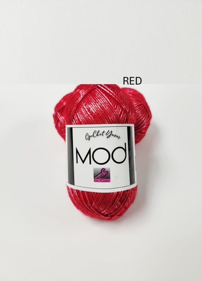 May include: A ball of bright red yarn with a metallic sheen. The yarn is wrapped in a white paper label featuring the text "Guchet Yarns MOD" and a small logo. The word "RED" is printed in black text above the yarn ball on the white background.