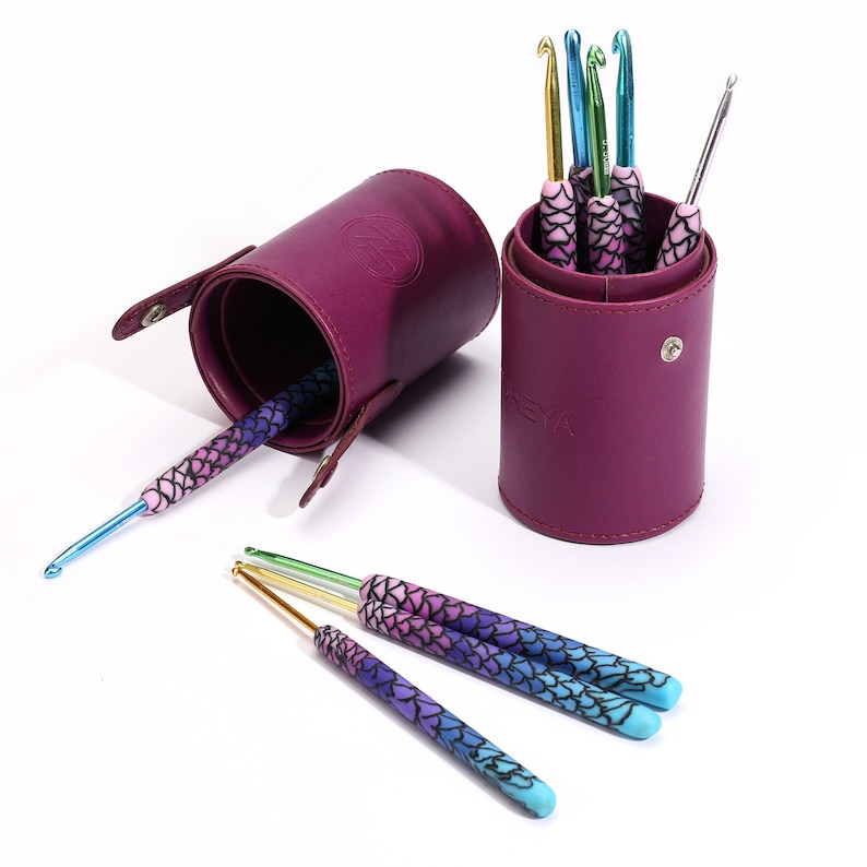 May include: A set of colorful crochet hooks with a mermaid scale design, stored in two purple leather cases. The cases have a zipper closure and a carrying handle.