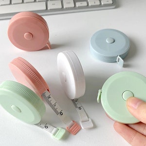 以下が含まれることがあります： ピンク、ライトブルー、ホワイト、ライトグリーンの4色のリトラクタブルテープメジャー。各テープメジャーは、ボタンでテープを巻き戻すことができる丸いプラスチックケースが付いています。