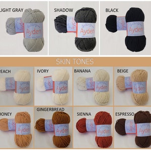 Op de afbeelding: Een verzameling Ayden-garen in diverse kleuren, waaronder lichtgrijs, schaduw en zwart. De afbeelding toont ook huidtinten zoals perzik, ivoor en banaan. Elk garen heeft een label met de merknaam en productdetails.