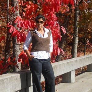 May include: A woman wearing a brown knitted vest over a white button-down shirt and dark blue jeans. She is standing in front of a wall with red leaves in the background.