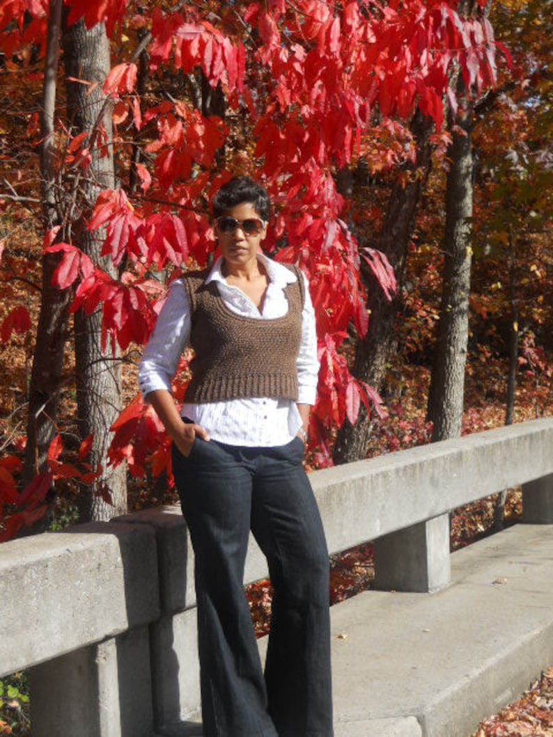 May include: A woman wearing a brown knitted vest over a white button-down shirt and dark blue jeans. She is standing in front of a wall with red leaves in the background.