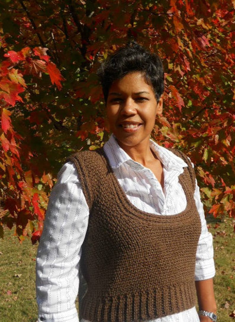 May include: A woman wearing a brown knitted vest over a white striped shirt. The vest has a scoop neck and is crocheted. The woman is standing in front of a tree with red and orange leaves.