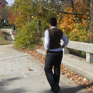 May include: A person wearing a brown sweater vest over a white shirt and black pants. The person is standing on a paved path with fallen leaves.