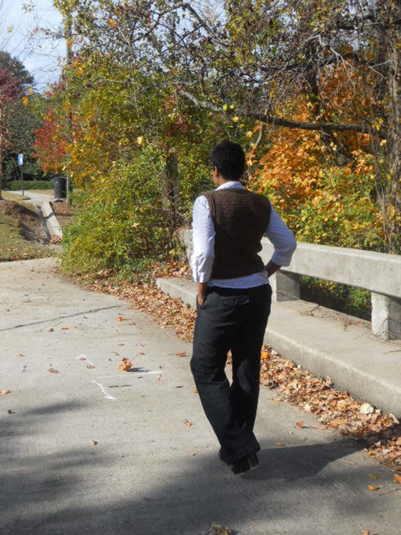 May include: A person wearing a brown sweater vest over a white shirt and black pants. The person is standing on a paved path with fallen leaves.
