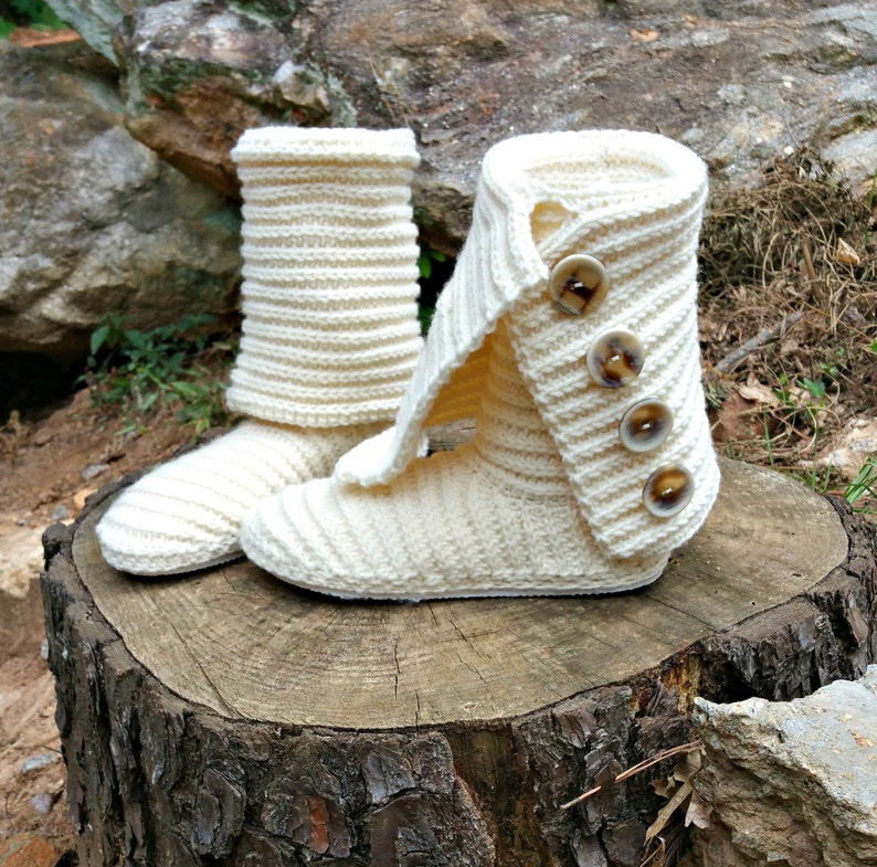 May include: A pair of white knitted slipper boots with fold-over cuffs and brown buttons. The boots are sitting on a tree stump.