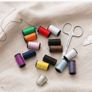 May include: A collection of colourful thread spools, a thimble, a pair of small scissors, and a needle on a white fabric surface.