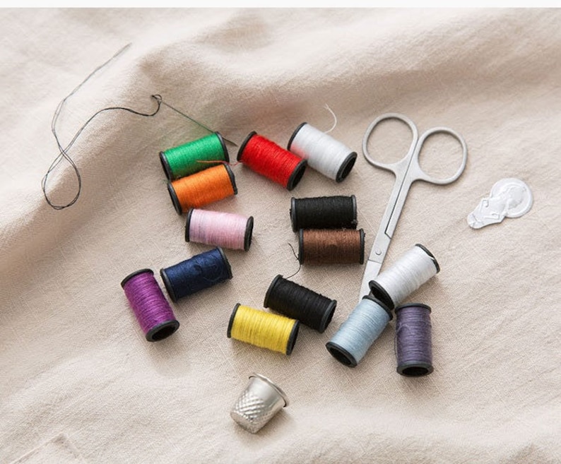 May include: A collection of colourful thread spools, a thimble, a pair of small scissors, and a needle on a white fabric surface.