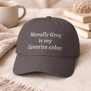 Może przedstawiać: Ciemnoszara czapka bejsbolówka z wyhaftowanym napisem "Morally Grey is my favorite color". Czapka ma zakrzywiony daszek i zaokrągloną górę. Kapelusz jest prezentowany na teksturowanej powierzchni.
