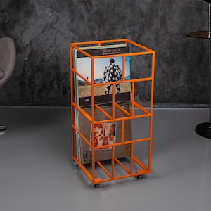 May include: An orange metal record storage unit on wheels. The unit has a rectangular frame with multiple compartments holding vinyl record albums. Album covers with band names and images are visible. The unit is on a gray floor.
