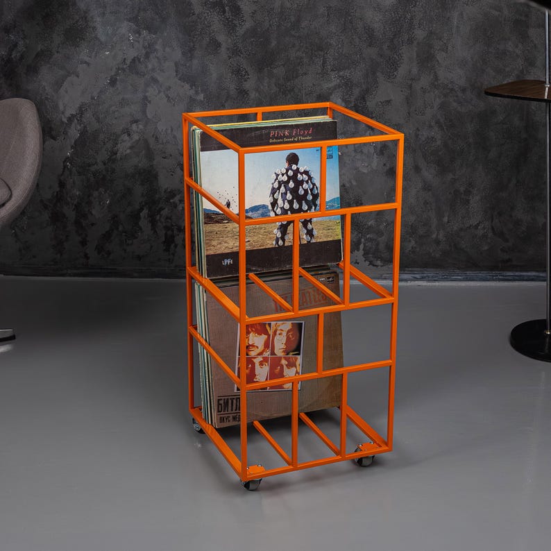 May include: An orange metal record storage unit on wheels. The unit has a rectangular frame with multiple compartments holding vinyl record albums. Album covers with band names and images are visible. The unit is on a gray floor.