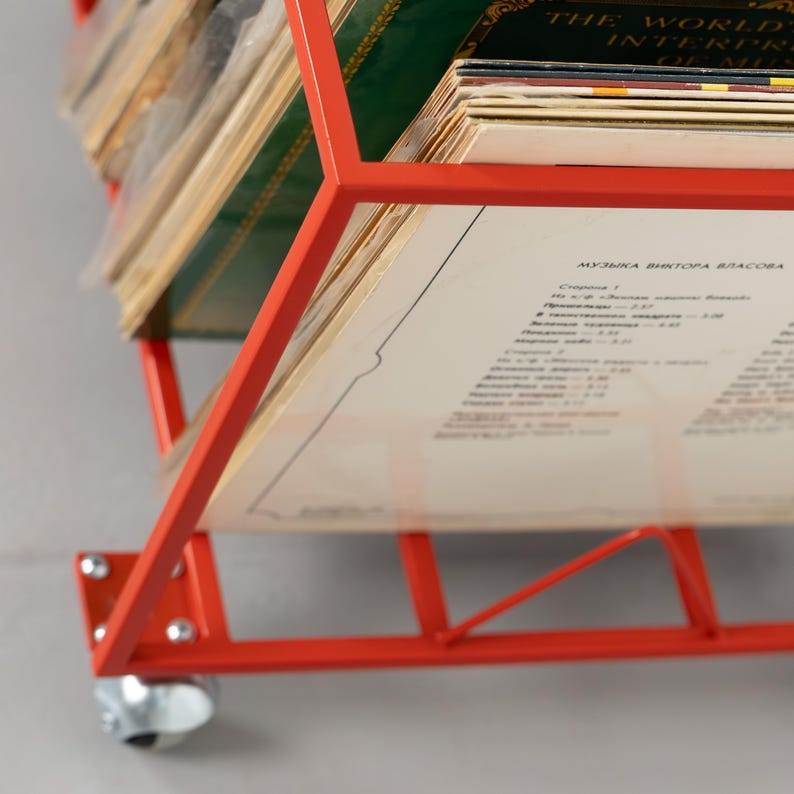 May include: A red metal record storage rack on wheels, holding a collection of vinyl records. The rack has a geometric design and is filled with album covers of various colors and designs. The text on one album is partially visible.