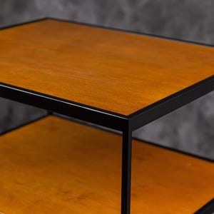 May include: A two-tier side table featuring a rectangular wooden top and a matching lower shelf, both finished in a warm medium-brown tone. The structure is supported by a thin, matte black metal frame that outlines each shelf edge.
