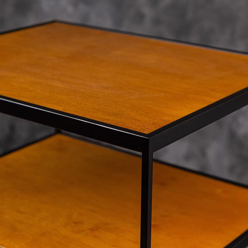 May include: A two-tier side table featuring a rectangular wooden top and a matching lower shelf, both finished in a warm medium-brown tone. The structure is supported by a thin, matte black metal frame that outlines each shelf edge.
