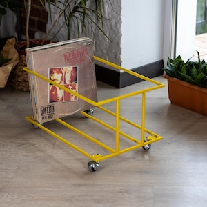 Può includere: Un portadischi in metallo giallo su ruote. Il supporto è rettangolare e contiene diversi dischi in vinile. La copertina dell'album visibile presenta i Beatles e il testo "A Taste of Honey".
