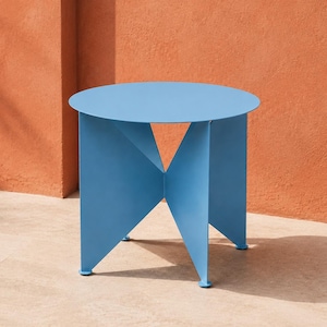 Peut inclure: Une table d'appoint ronde en métal bleu clair dotée d'un plateau circulaire plat reposant sur une base composée de quatre panneaux verticaux triangulaires entrecroisés formant une structure géométrique en croix avec de petits pieds ronds.