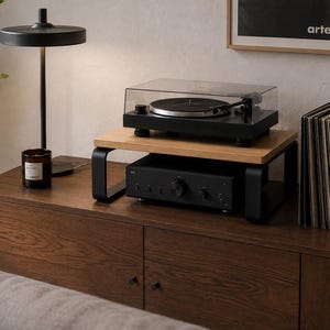 May include: A wooden monitor stand with black metal legs supporting a black turntable with a clear dust cover. Beneath the stand sits a black audio amplifier. The unit is placed on a dark wood cabinet next to a stack of vinyl records.