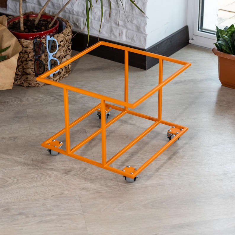 Op de afbeelding: Een oranje metalen plantenstandaard met een rechthoekig frame en vier kleine wielen. De standaard heeft een ontwerp met twee niveaus, met een boven- en onderframe verbonden door verticale steunen. De standaard is ontworpen om planten te bevatten.