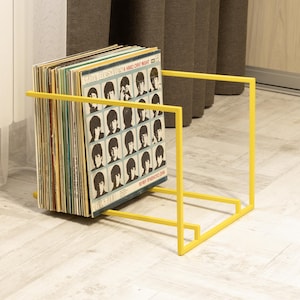 May include: A bright yellow metal record holder with a stack of vinyl records. The album cover visible features black and white portraits of The Beatles. The holder has a minimalist, geometric design.