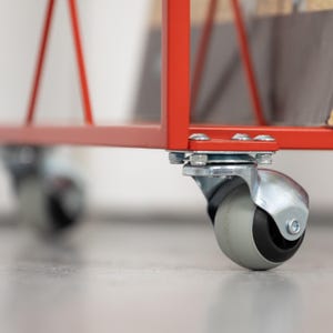 May include: Close-up of a red metal cart frame with a caster wheel. The wheel has a gray tire and a silver metal housing. The cart frame is attached to the wheel with bolts and nuts. The background is blurred.