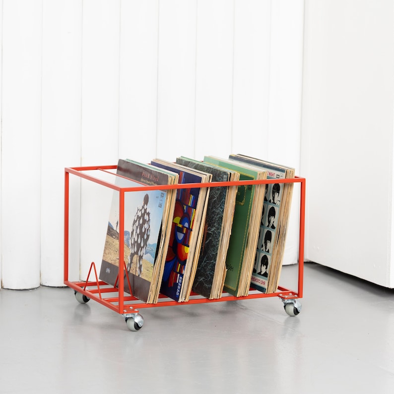 May include: A red metal record storage cart on wheels, holding several vinyl record albums. The cart has a rectangular frame and a sloped base to support the records. The albums have various colorful covers.