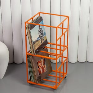 May include: An orange metal record storage rack on wheels. The rack has a rectangular frame with multiple compartments holding vinyl records. The records have album art visible. The rack is on a gray floor with a white background.