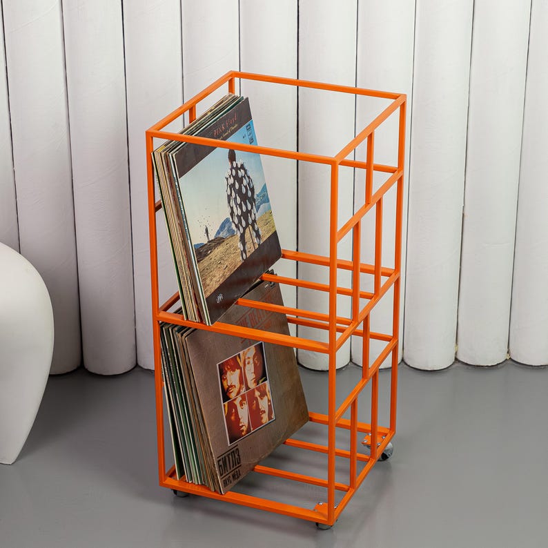 May include: An orange metal record storage rack on wheels. The rack has a rectangular frame with multiple compartments holding vinyl records. The records have album art visible. The rack is on a gray floor with a white background.