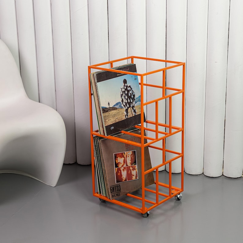 May include: An orange metal record storage rack on wheels. The rack is filled with vinyl records. The rack has a rectangular shape with a grid-like design. The records have album art visible.