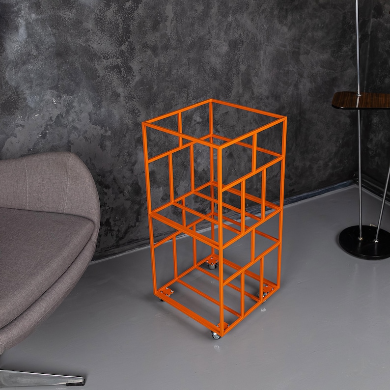 May include: An orange metal storage unit with a geometric design. The unit has a rectangular shape with multiple open compartments and sits on small wheels for mobility. The frame is a bright orange color.