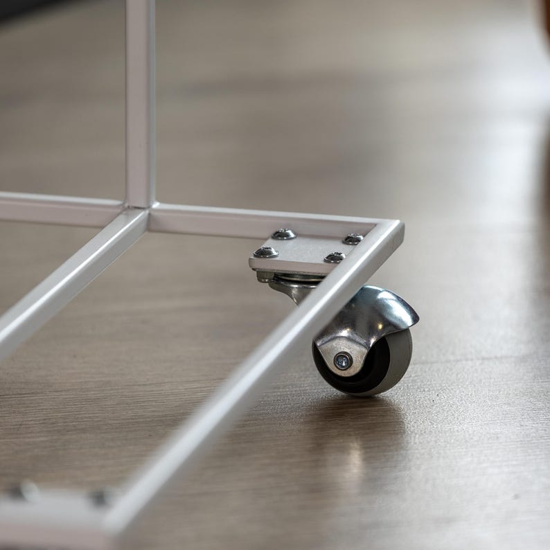 Op de afbeelding: Detail van een witte metalen hoek met een klein, grijs zwenkwiel. Het wiel is bevestigd aan een zilveren metalen plaat met schroeven. Het frame staat op een lichtbruine houten vloer. Het wiel heeft een diameter van ongeveer 5 cm.