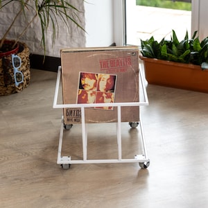 Puede incluir: Un soporte de discos de metal blanco sobre ruedas, que muestra una portada de álbum vintage de The Beatles. La portada del álbum presenta cuatro retratos de miembros de la banda. El soporte es rectangular con un diseño de rejilla.