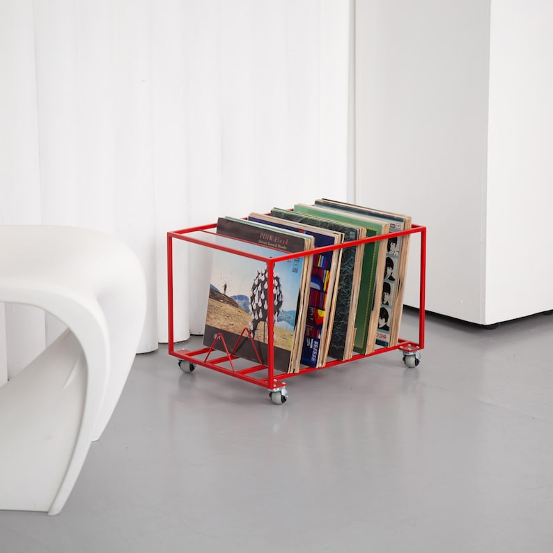 May include: A red metal record storage cart on wheels, holding several vinyl record albums. The cart has a rectangular frame and a wire rack to hold the records upright. The albums have various colorful covers.