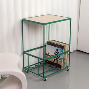 Op de afbeelding: Een groene metalen bijzettafel met een licht houten blad en een onderste plank voor vinylplaten. De tafel heeft een rechthoekig frame en staat op vier kleine wielen. Een stapel vinylplaten is zichtbaar op de plank.