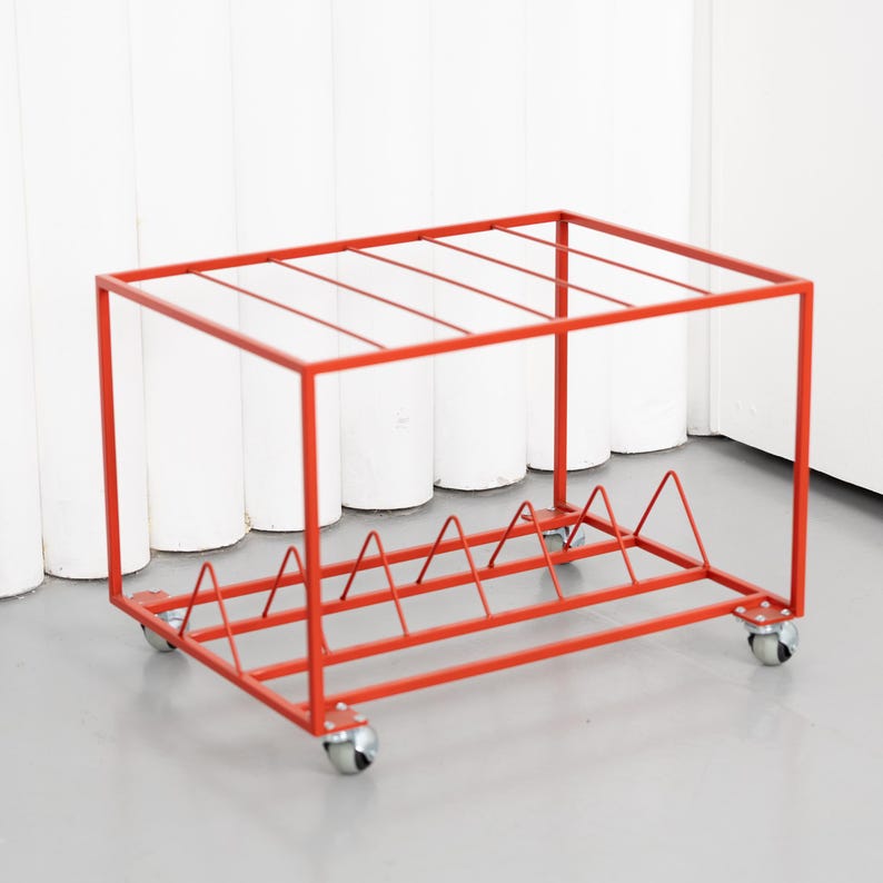 May include: A red metal storage rack on wheels. The rack has a rectangular frame with a series of horizontal bars on top and a triangular design at the bottom. The rack is designed to hold items and has four small wheels.
