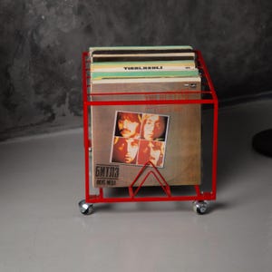 May include: A red metal record storage cart on wheels, holding several vinyl record albums. The cart has a rectangular frame and a triangular support. One album cover features a band with four members.