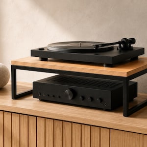 May include: A black turntable sits on top of a rectangular wooden monitor stand with a black metal frame. Beneath the stand, a black audio amplifier with a central volume knob and several smaller control buttons is positioned on a wooden surface.