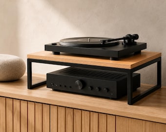 May include: A black turntable sits on top of a rectangular wooden monitor stand with a black metal frame. Beneath the stand, a black audio amplifier with a central volume knob and several smaller control buttons is positioned on a wooden surface.